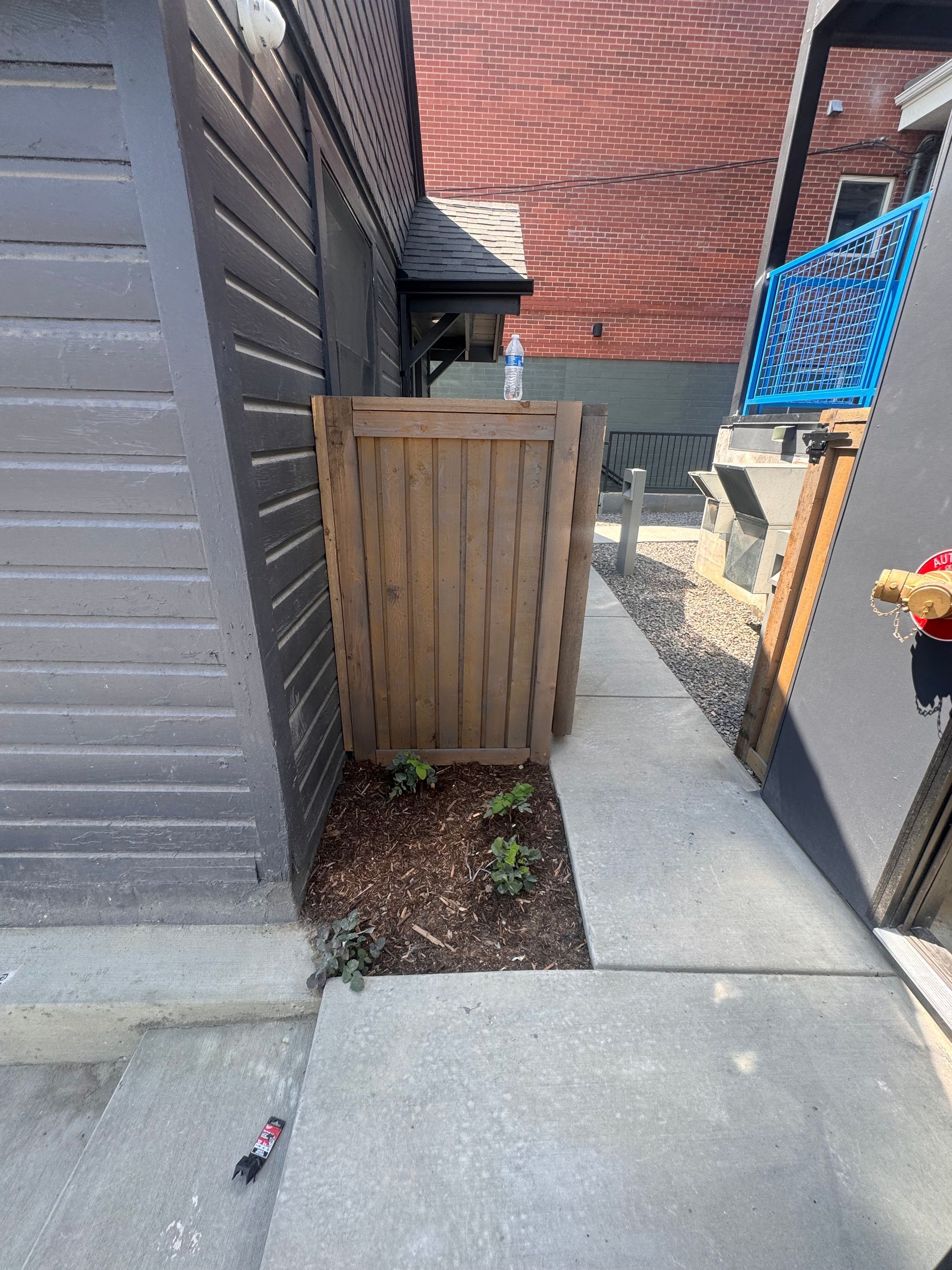 A narrow walkway bordered by a brown wooden fence, a gray building, and a brick wall. Small plants at base of the fence.
