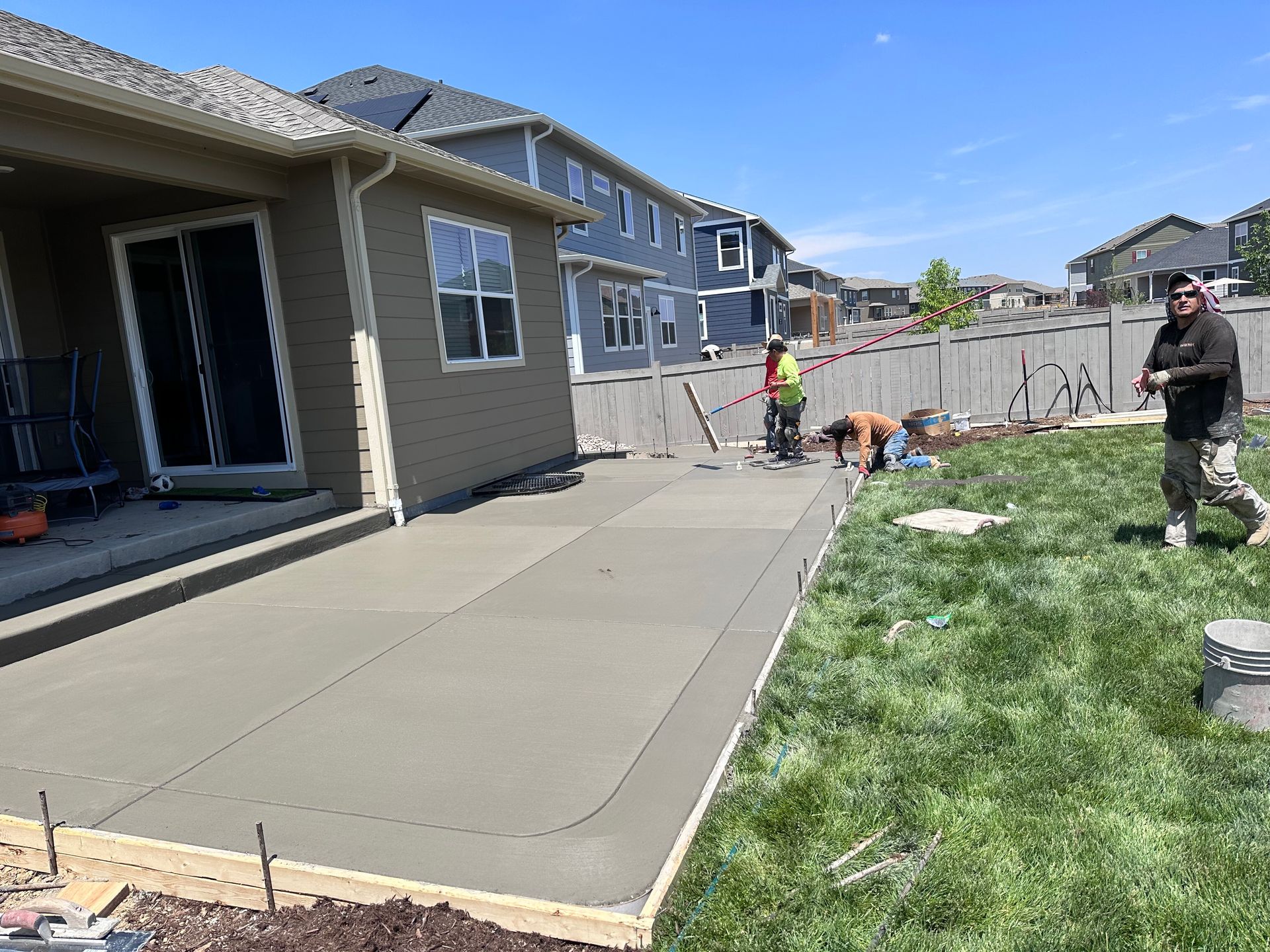 Workers pouring concrete patio in a backyard with a house, grass, and fence. Bright sunny day.