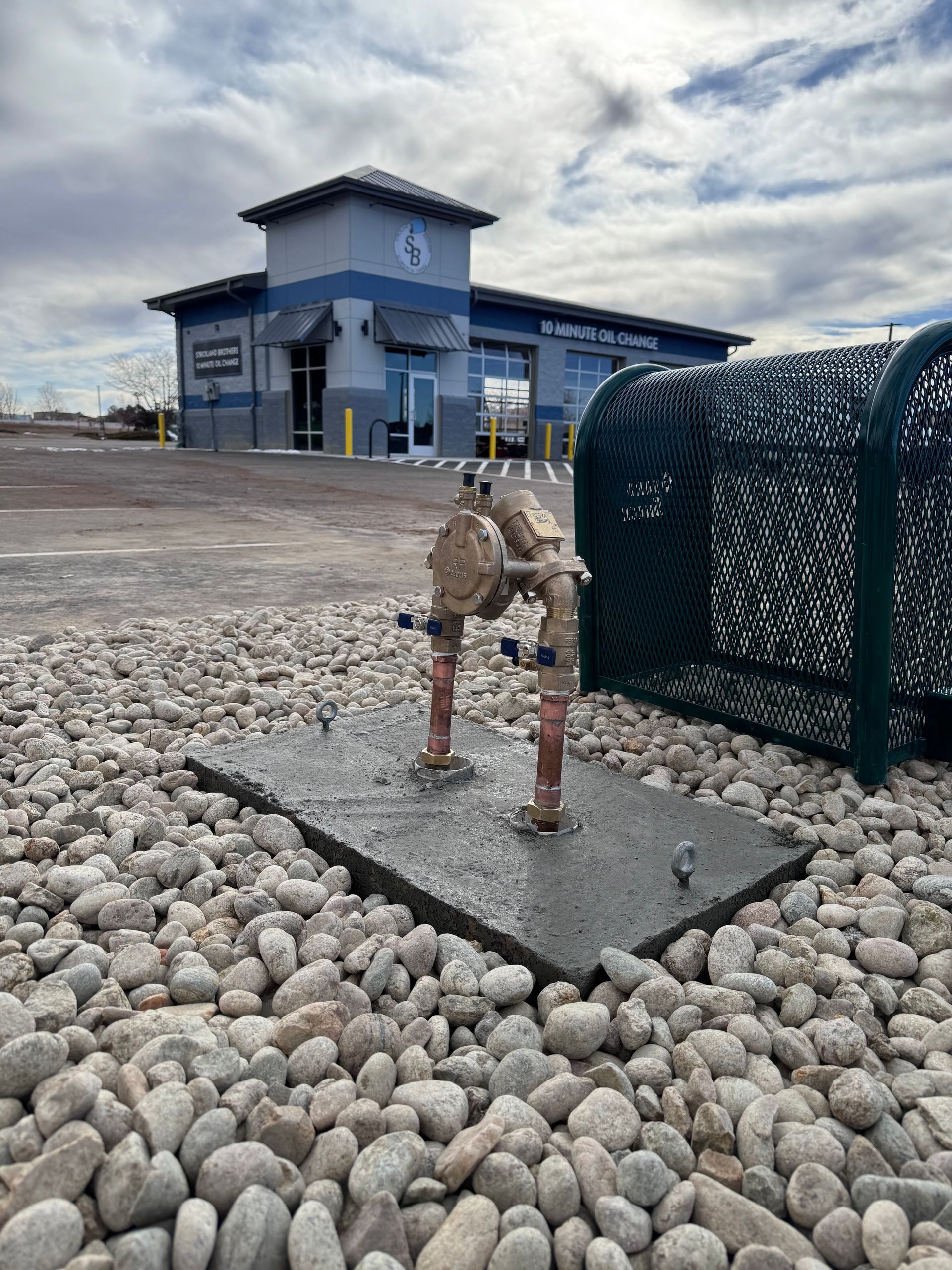 Water meter with pipes on concrete base in front of a blue car wash building with gravel.