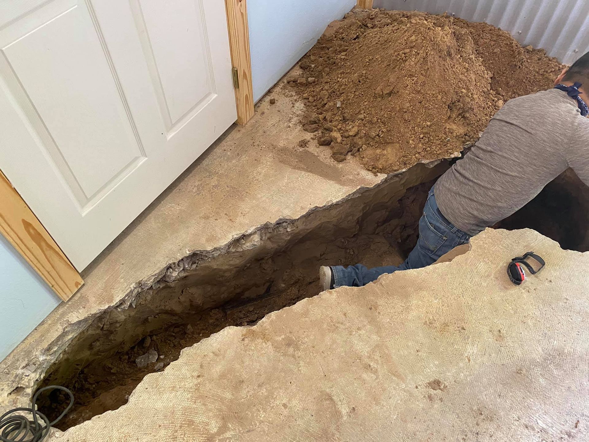 A person in a gray shirt and blue jeans reaches into a deep, long trench cut into a concrete floor inside a building.