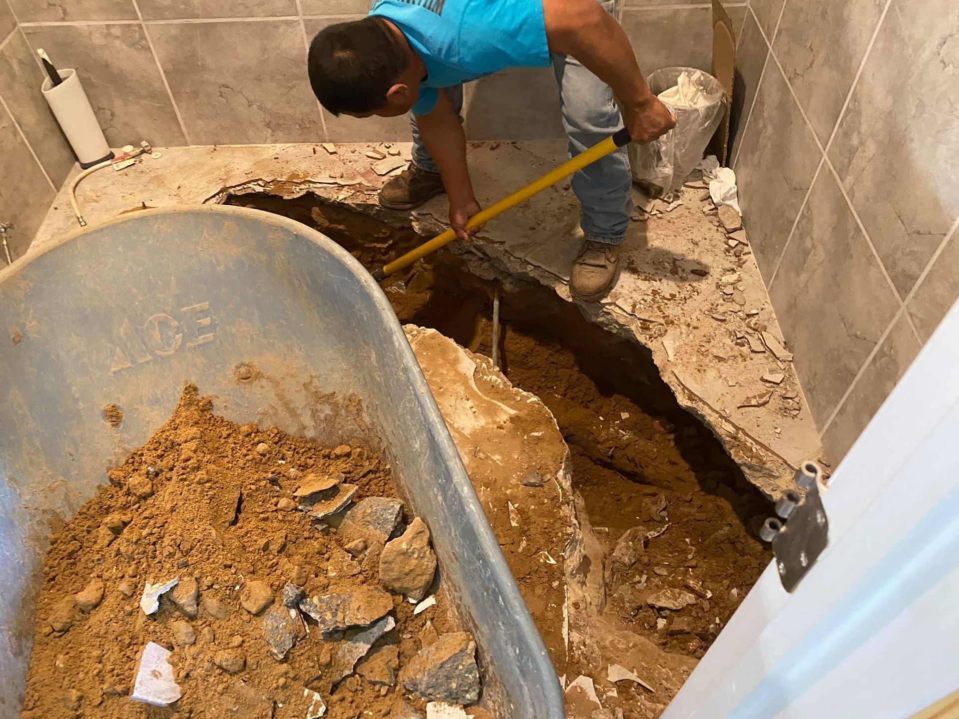 A worker in a blue shirt uses a yellow-handled tool to dig into the ground inside a bathroom with exposed soil and rubble.