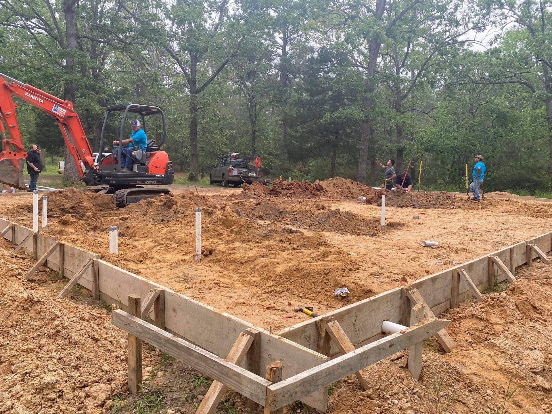 A construction site showing workers and an orange excavator preparing a foundation with wooden forms in a wooded area.