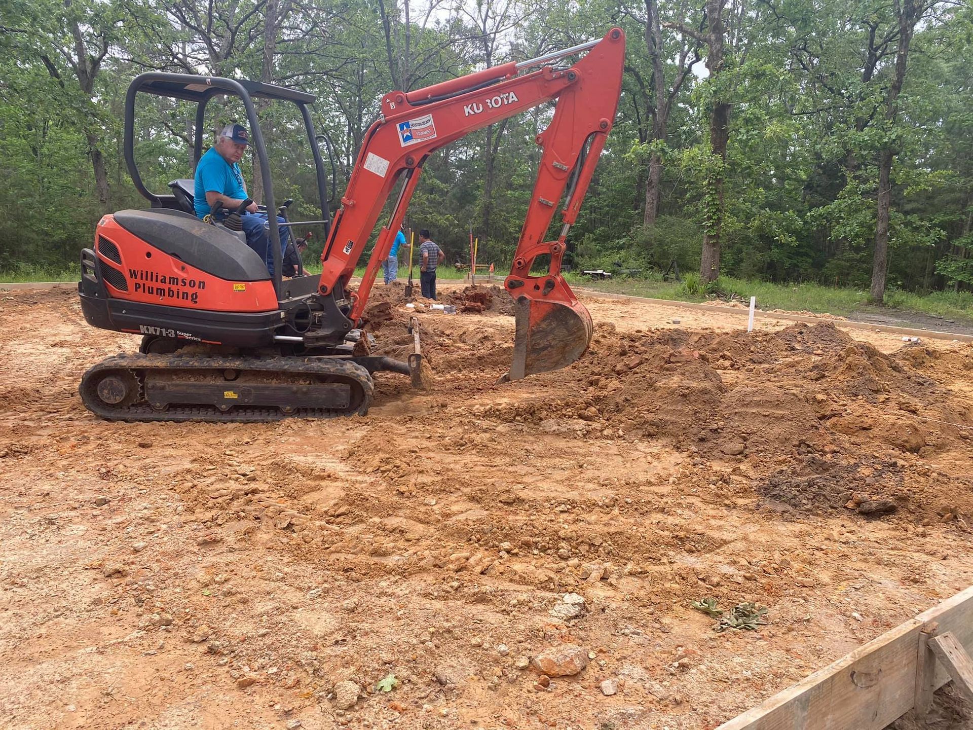 A person operating an orange compact excavator to dig in a dirt construction site surrounded by trees.