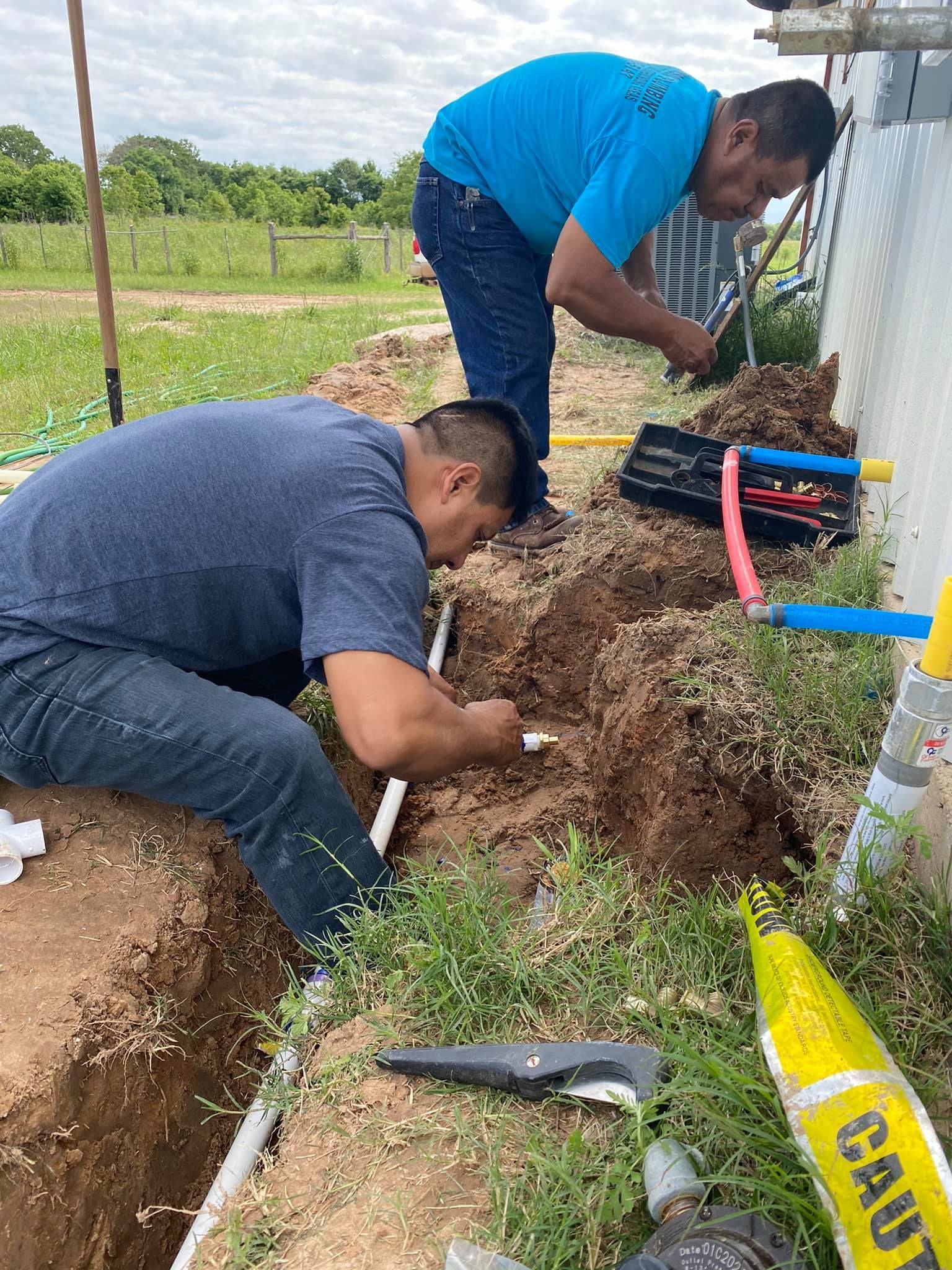 Two people work in a trench next to a building, connecting blue and white pipes near a black electrical box.