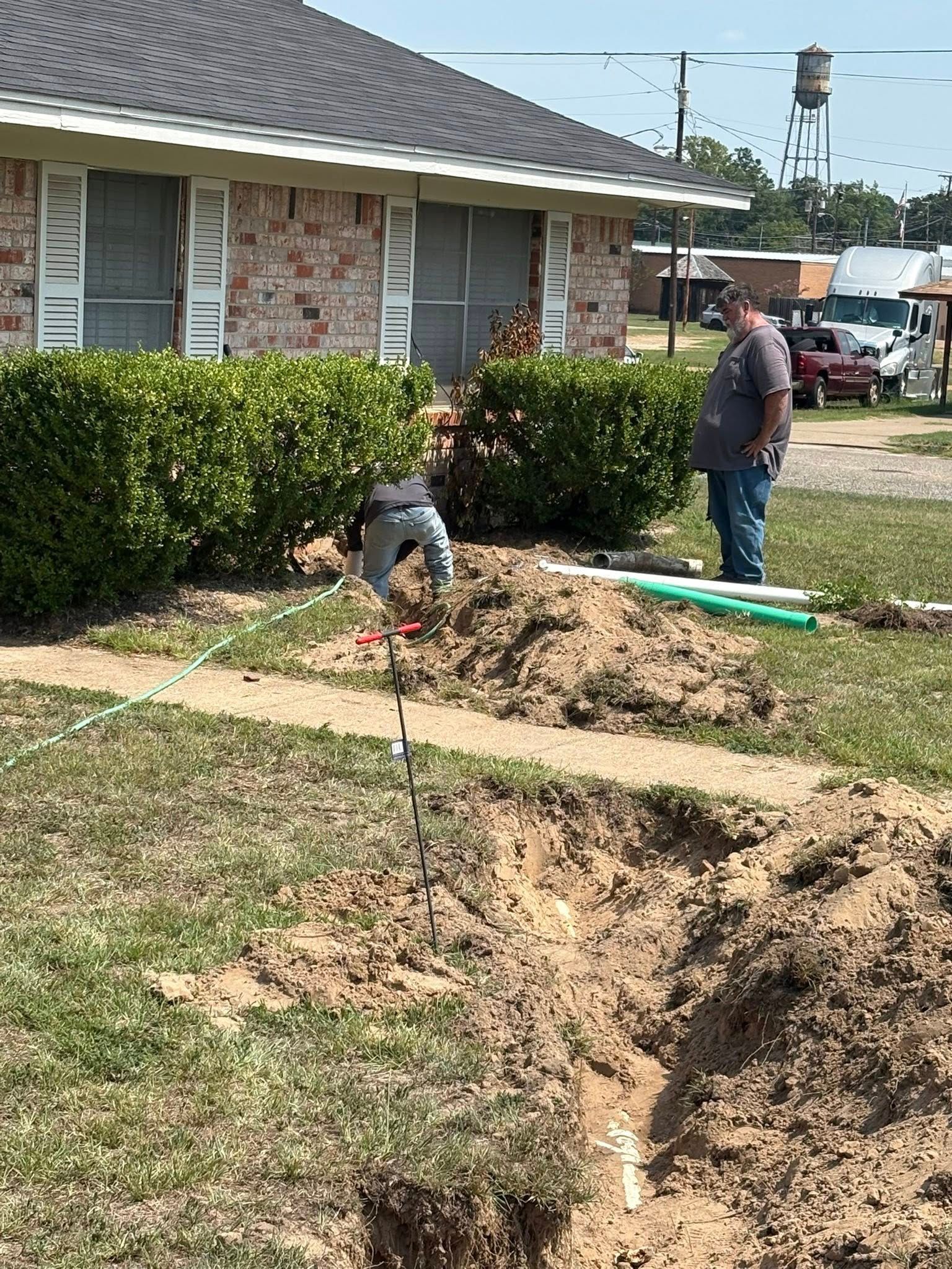 A person stands next to a trench in a yard while another person works in the dirt near a brick house.