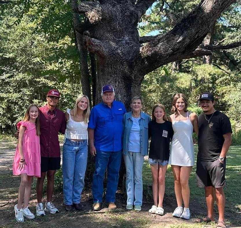 A group of eight people standing side-by-side in front of a large tree in a sunlit wooded area.