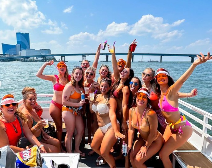 Group of women in swimwear on a boat, smiling with arms raised, Atlantic City backdrop.