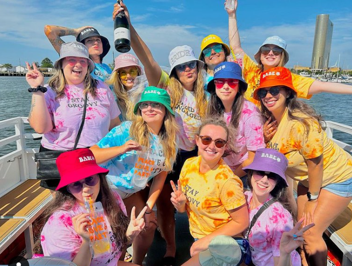 Group of women on a boat celebrating, wearing colorful tie-dye shirts and bucket hats.