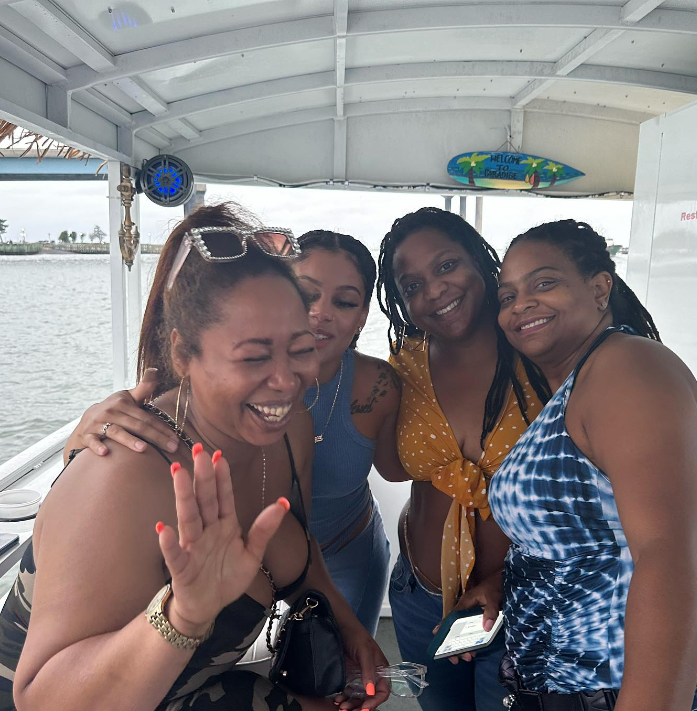 Four smiling people on a boat, waving, posing. Sunny day, water in background.