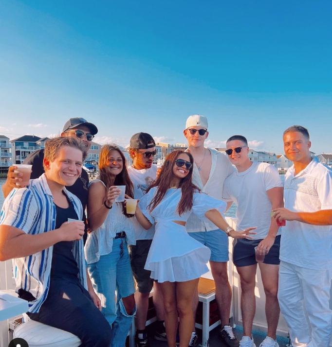 Group of people on a boat deck smiling, with blue sky and coastal houses in the background.