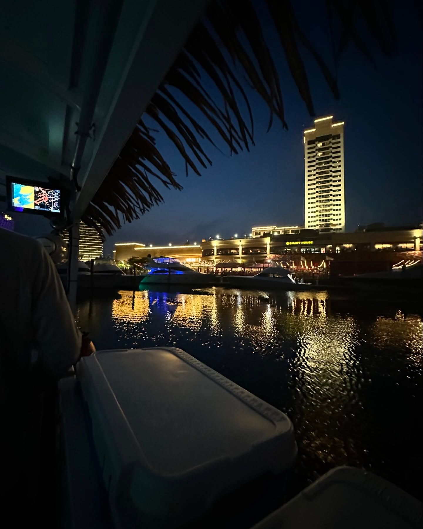 Night view of a city waterfront with a tall lit building reflected in dark water. Boat interior visible.