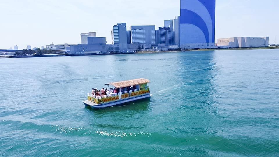 Boat with passengers on water, with city buildings in background, on a sunny day.