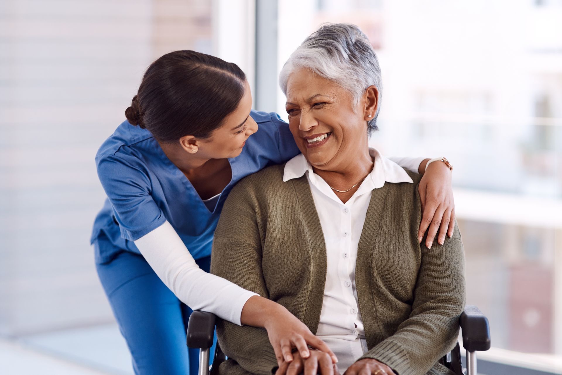 Nurse in blue scrubs with arm around smiling person in a wheelchair