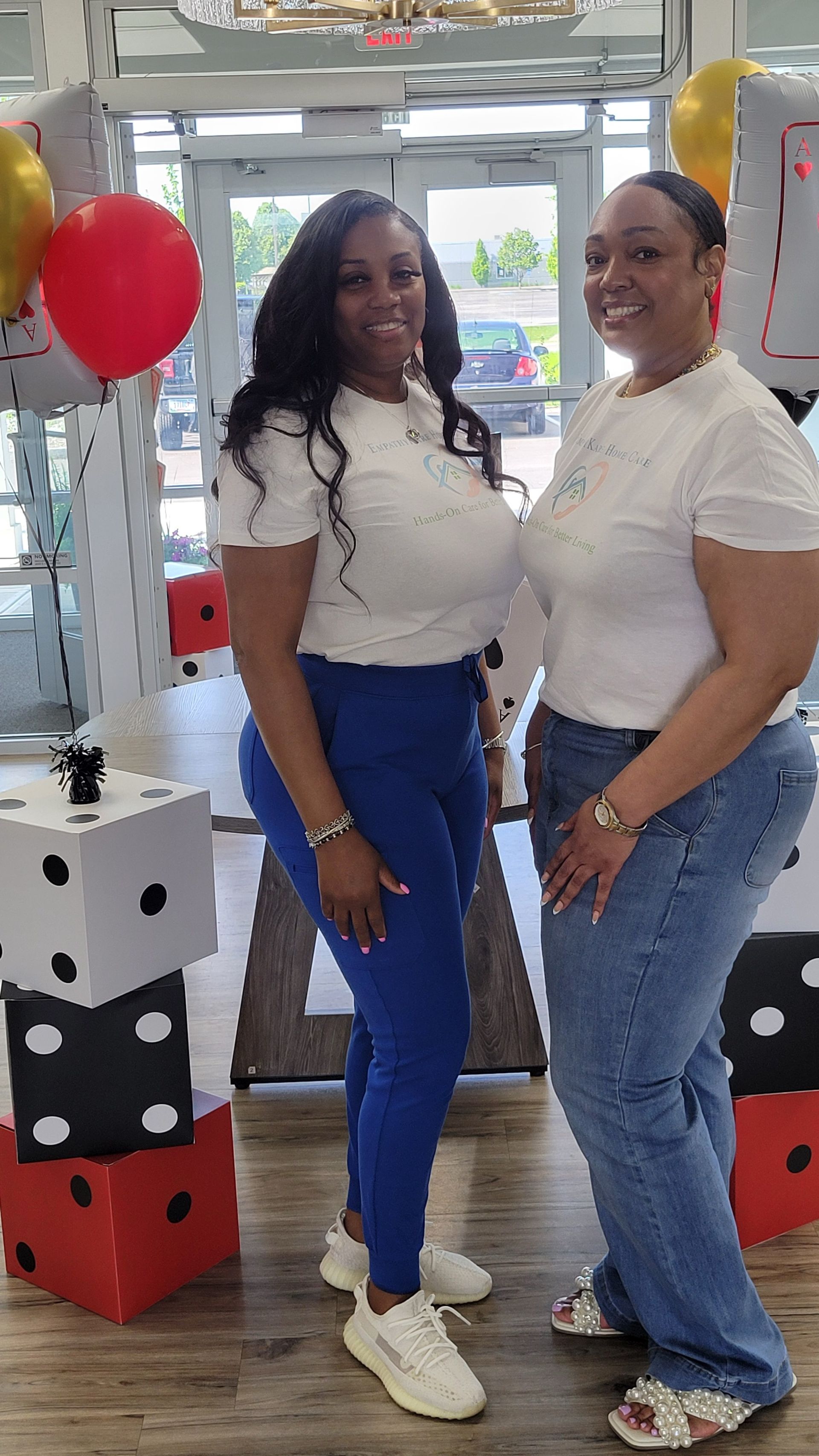 Two women in white shirts pose by dice decorations and balloons