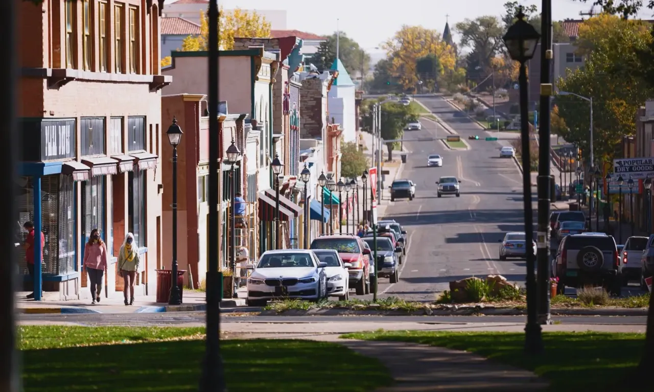 A street view of a small town with storefronts on the left, parked cars, and vehicles driving down the road.