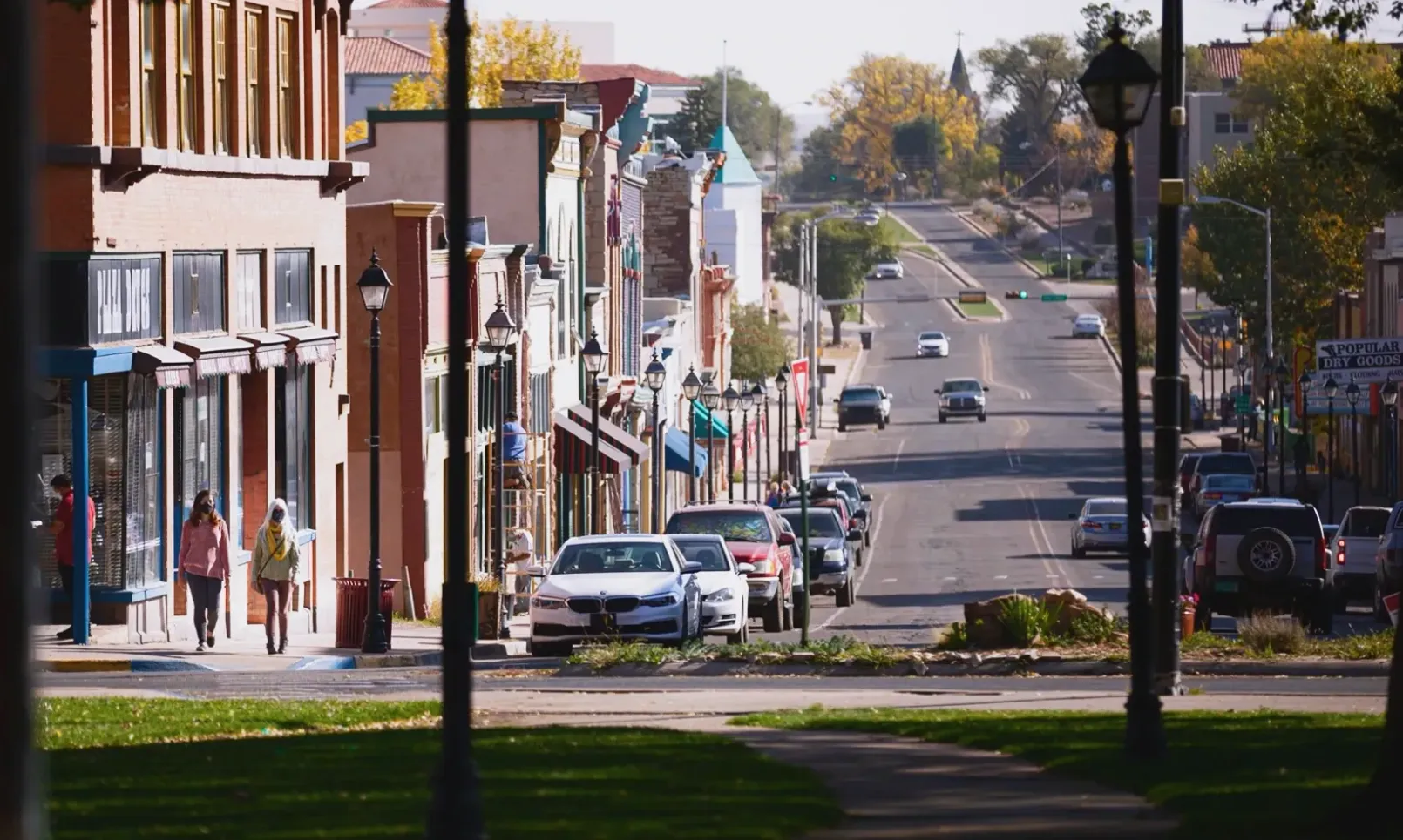 A street view of a small town with storefronts on the left, parked cars, and vehicles driving down the road.
