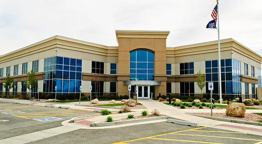 A two-story commercial building with tan siding and blue tinted windows, featuring a central entrance and a flagpole.