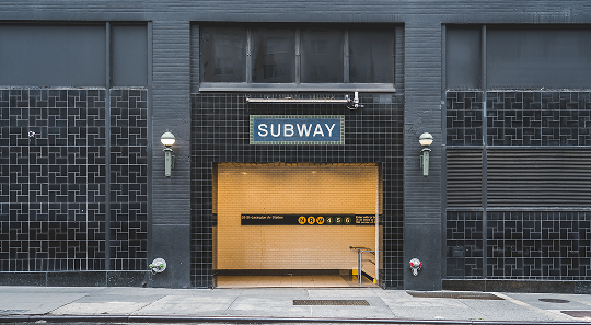 A subway entrance with a blue sign, black tiled walls, and bright interior lighting, situated on a city sidewalk.