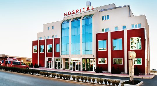 A modern multi-story hospital building with red accents, a glass central facade, and a row of shrubs in the foreground.
