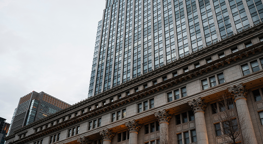 A tall, glass-faced skyscraper rises behind a historic stone building featuring prominent columns and classic architecture.