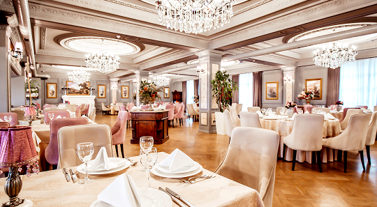 An elegant restaurant dining room featuring crystal chandeliers, light-colored upholstered chairs, and set tables.