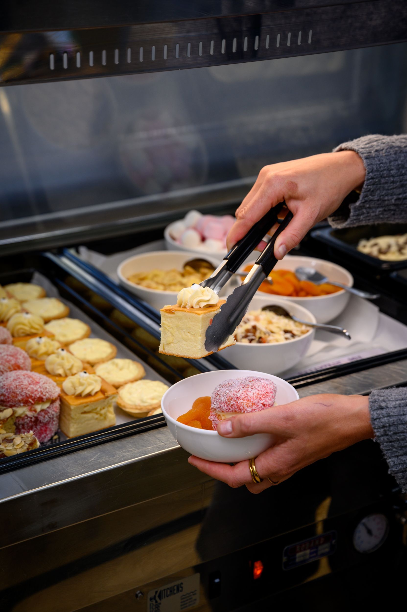 Person using tongs to serve cake from a buffet display at Collie Hills Village, other food items visible.