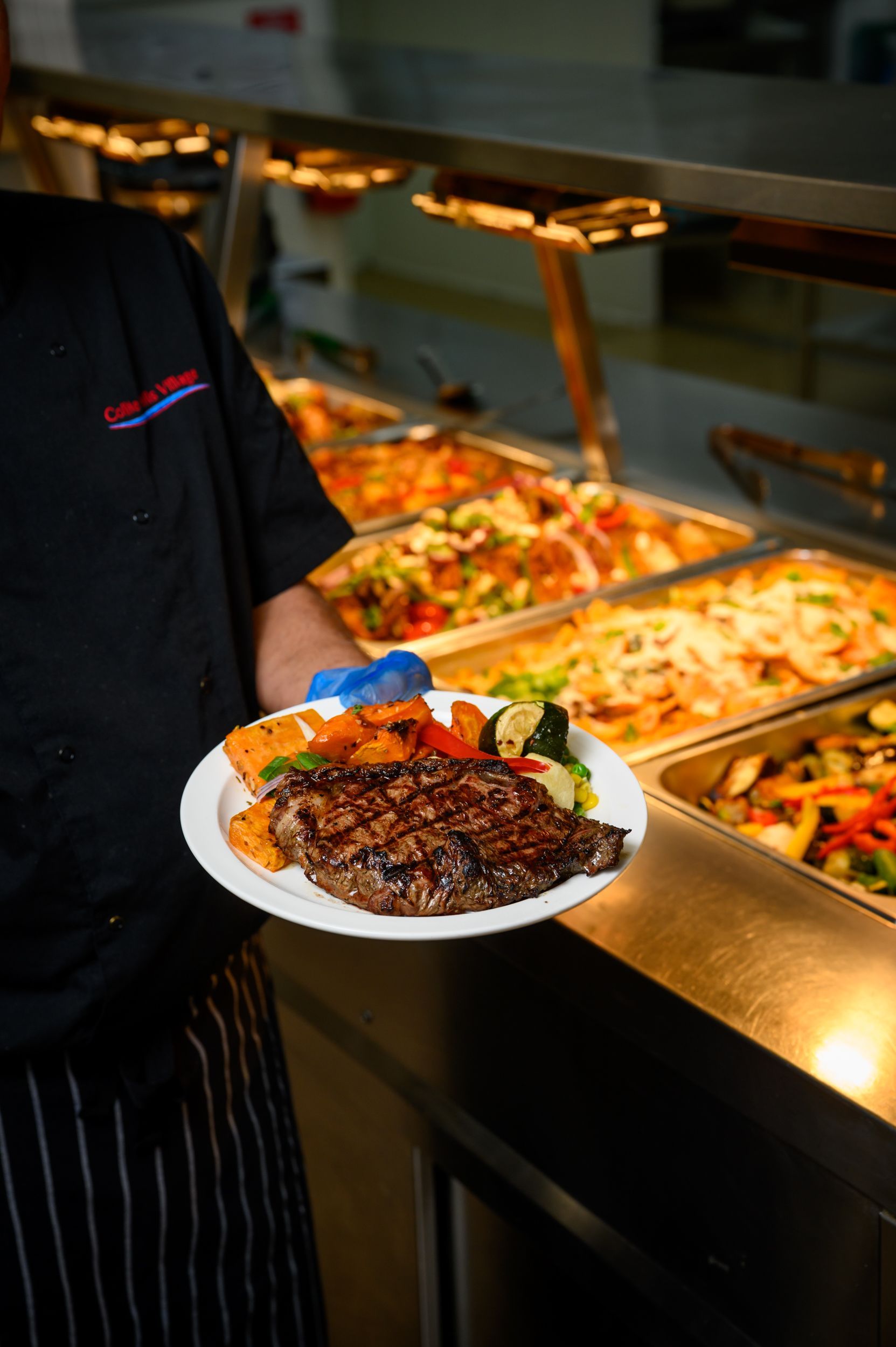 Chef holding plate with steak, sweet potatoes, and vegetables, in front of a buffet.