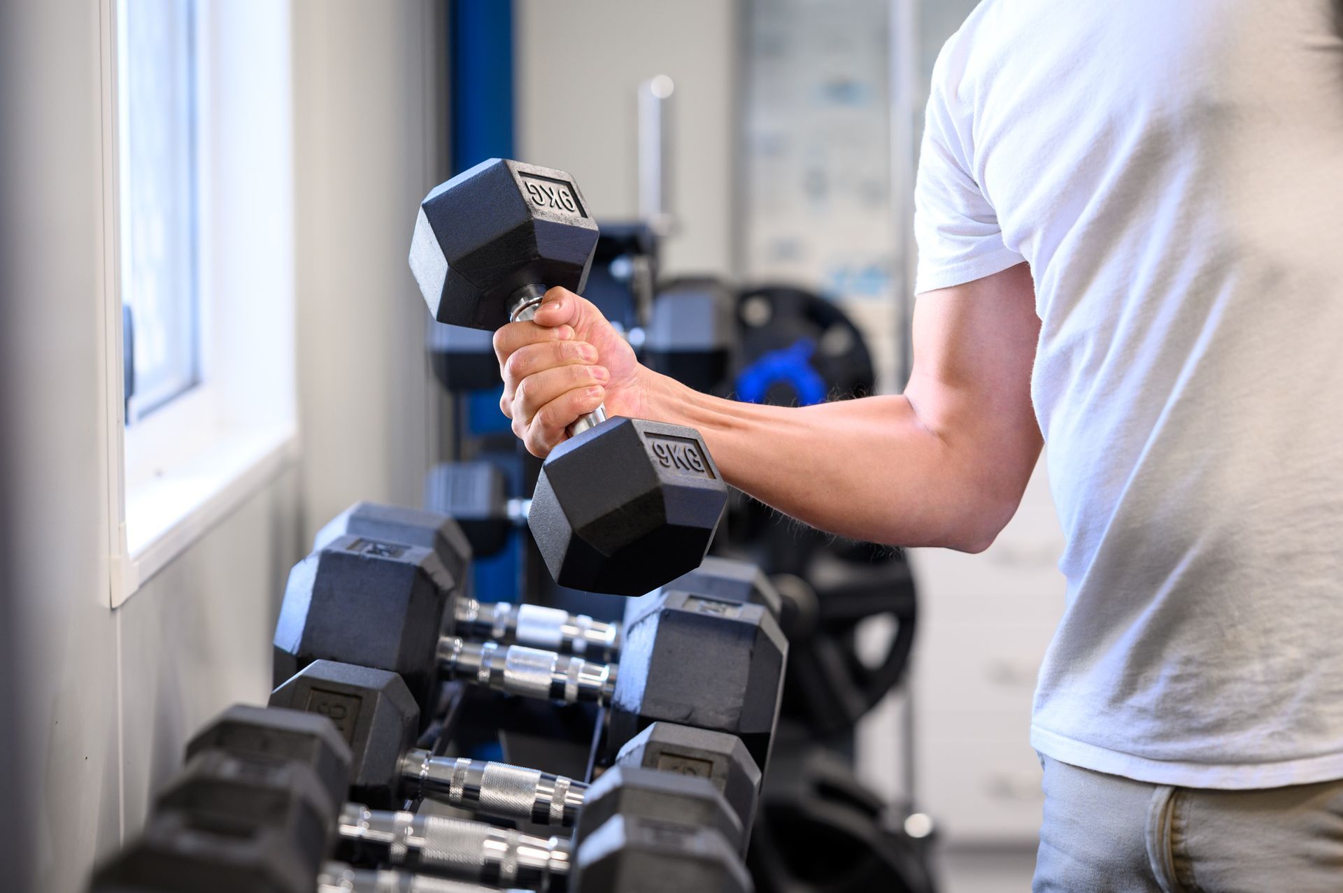 Man lifting a black dumbbell in a gym with a rack of weights.