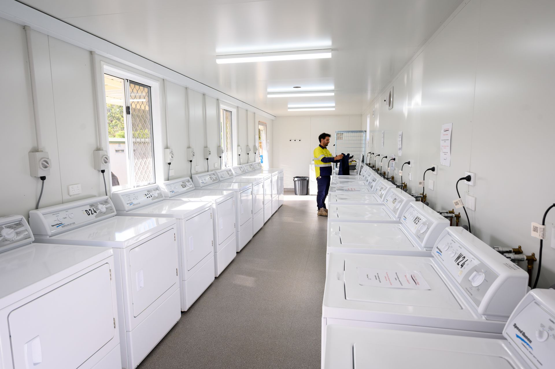 Laundry room at Collie Hills Village with rows of white washing machines. Person stands by machines. Bright interior.