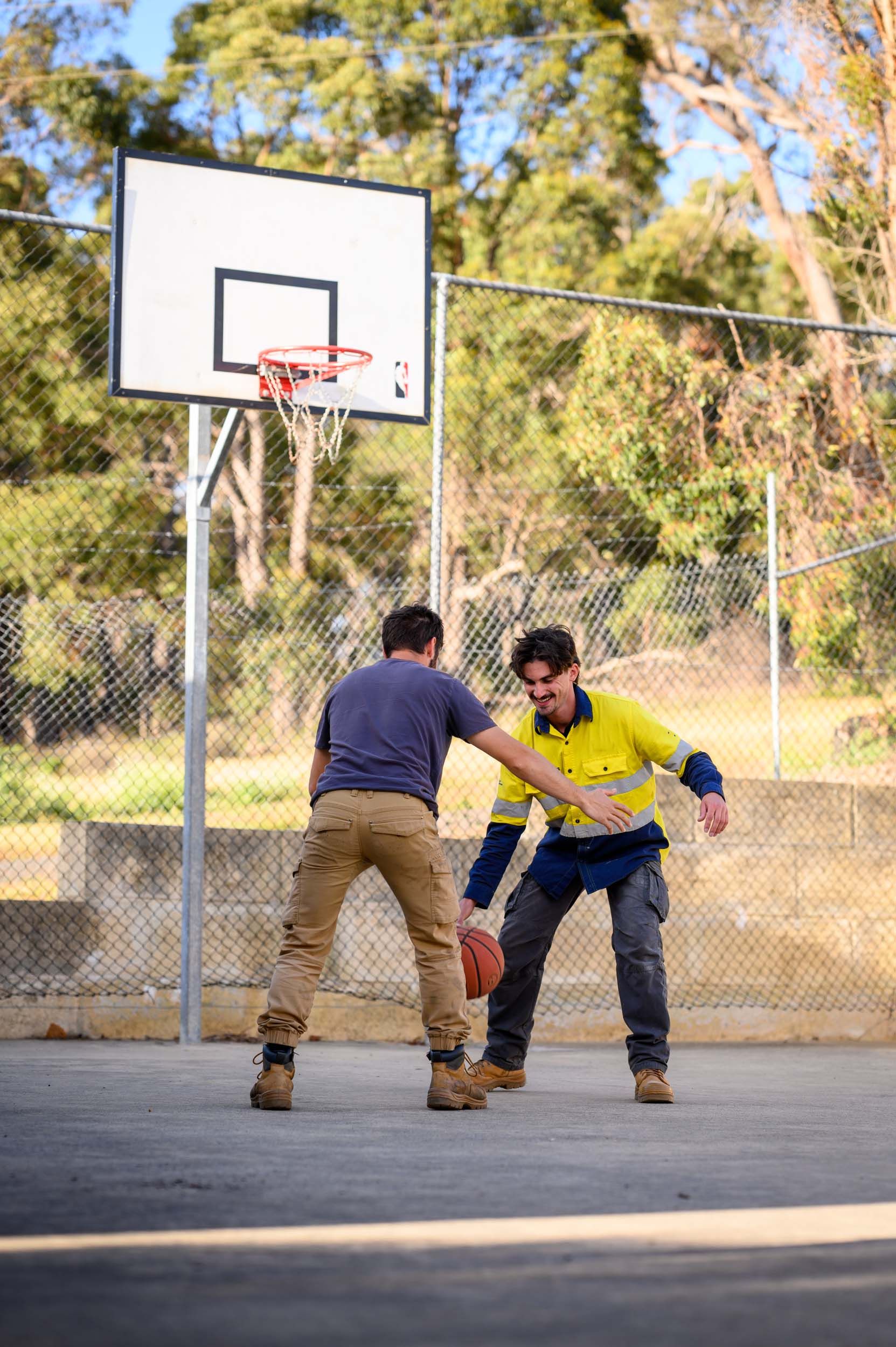 Two people playing basketball outdoors. One dribbles, other defends. Hoop, fence, trees visible.