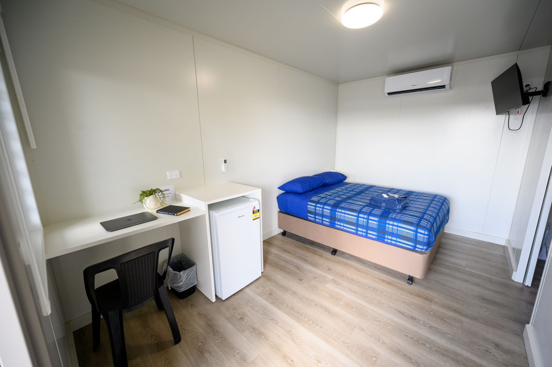 A small, simple bedroom with a bed, desk, chair, mini-fridge, and mounted TV.
