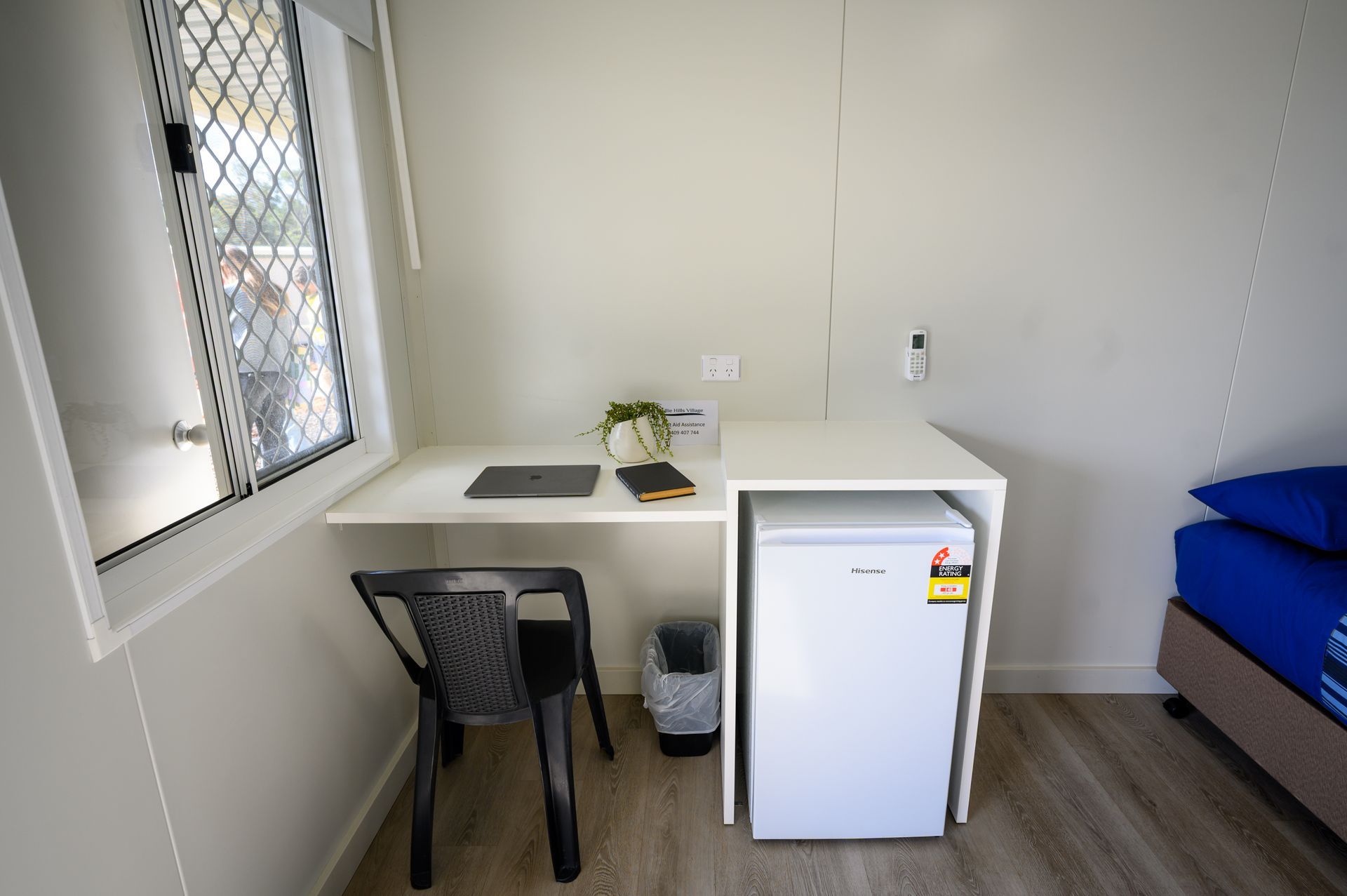Small white room with desk, chair, laptop, and mini-fridge near window and bed.