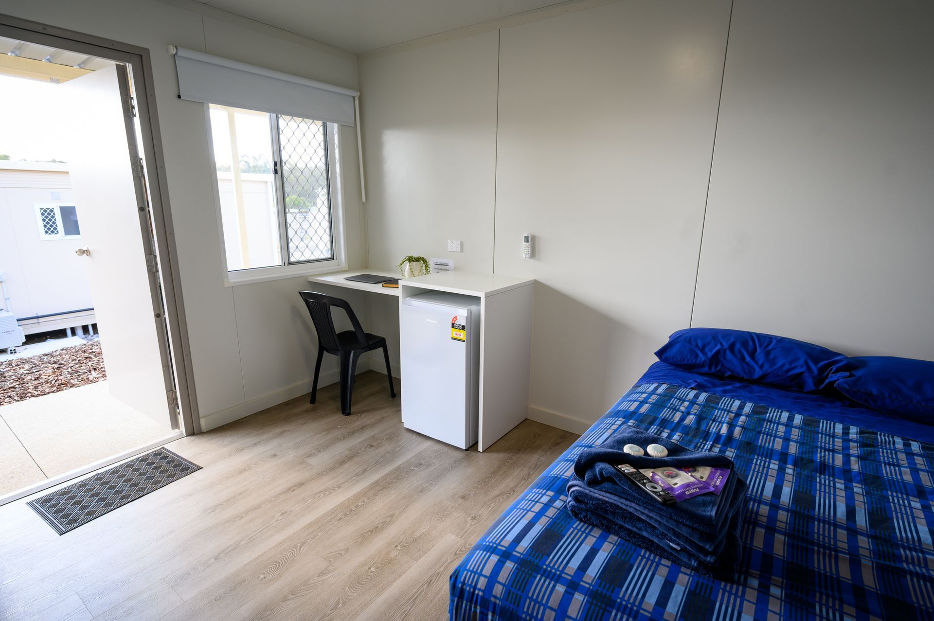 Small room with bed, desk, and open doorway to patio. Blue bedspread and white walls.