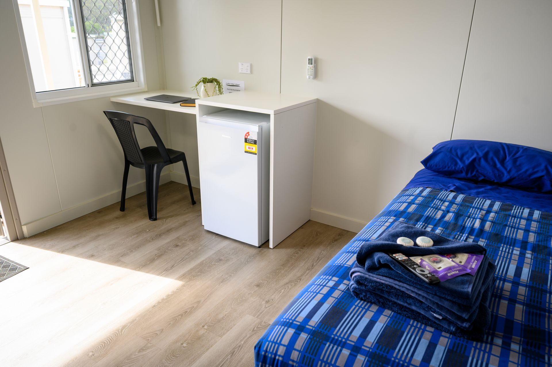 Small room with a single bed, desk with chair, mini-fridge, and window. Blue and white patterned bedspread.