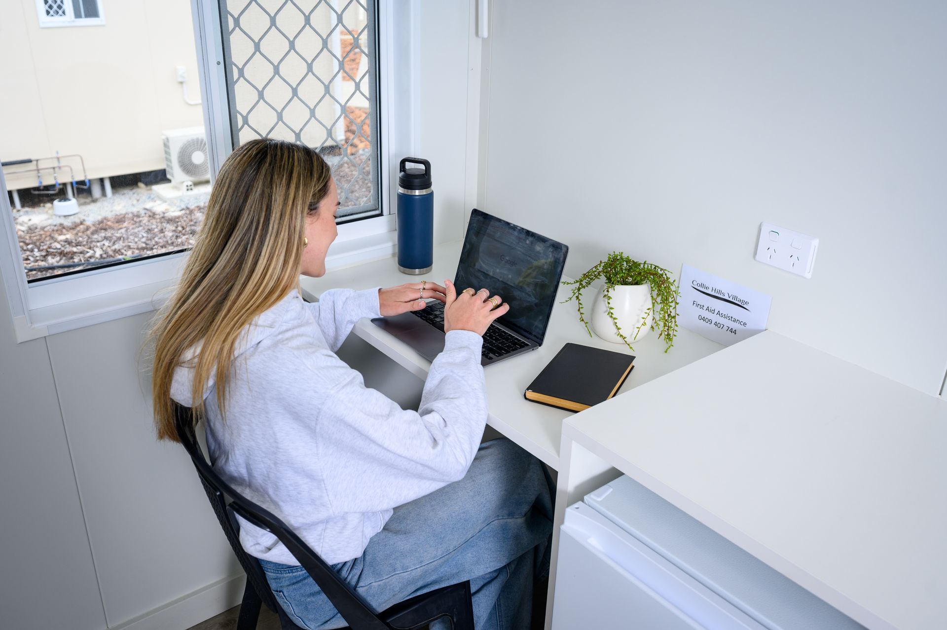 Woman at desk typing on laptop in a small, well-lit room.