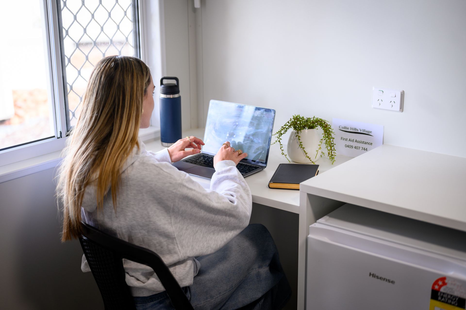 Woman working on laptop at a desk by a window; water bottle, plant, and notebook nearby.