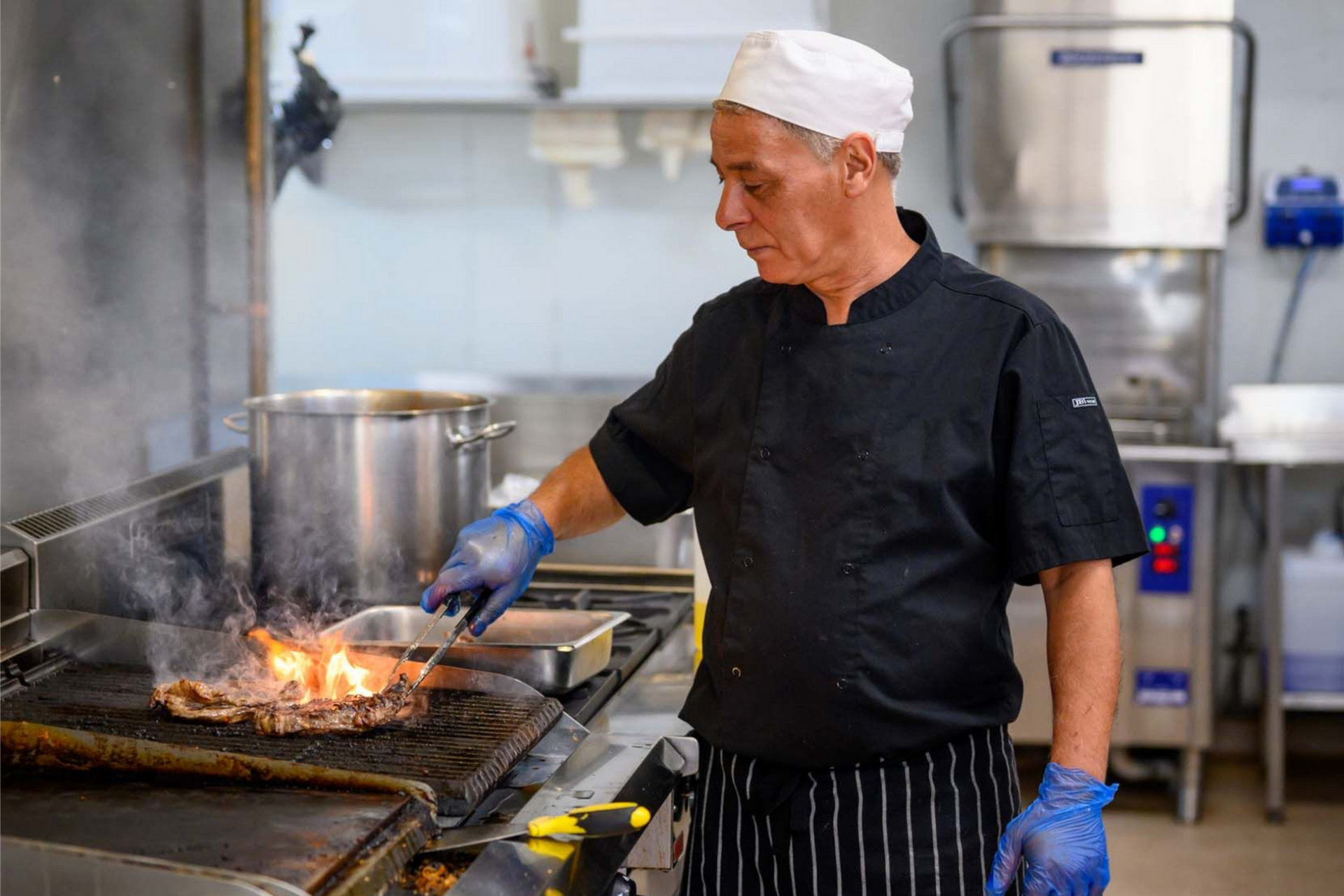 Chef grilling steak in commercial kitchen. Flames visible.