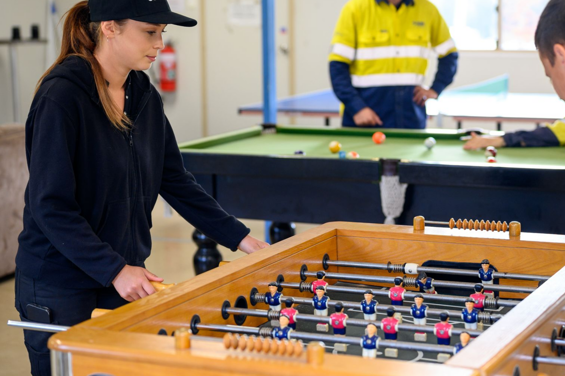 Woman playing foosball, wearing a black cap and jacket, with others playing pool in the background.
