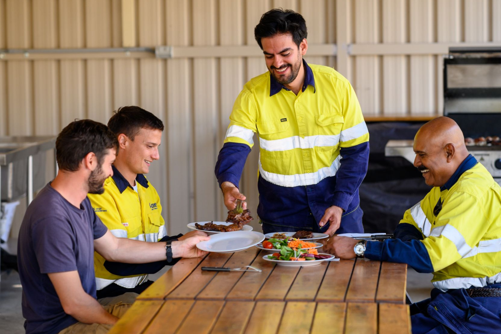 Four workers in reflective vests share food around a wooden table in an outdoor shelter at Collie Hills Village.