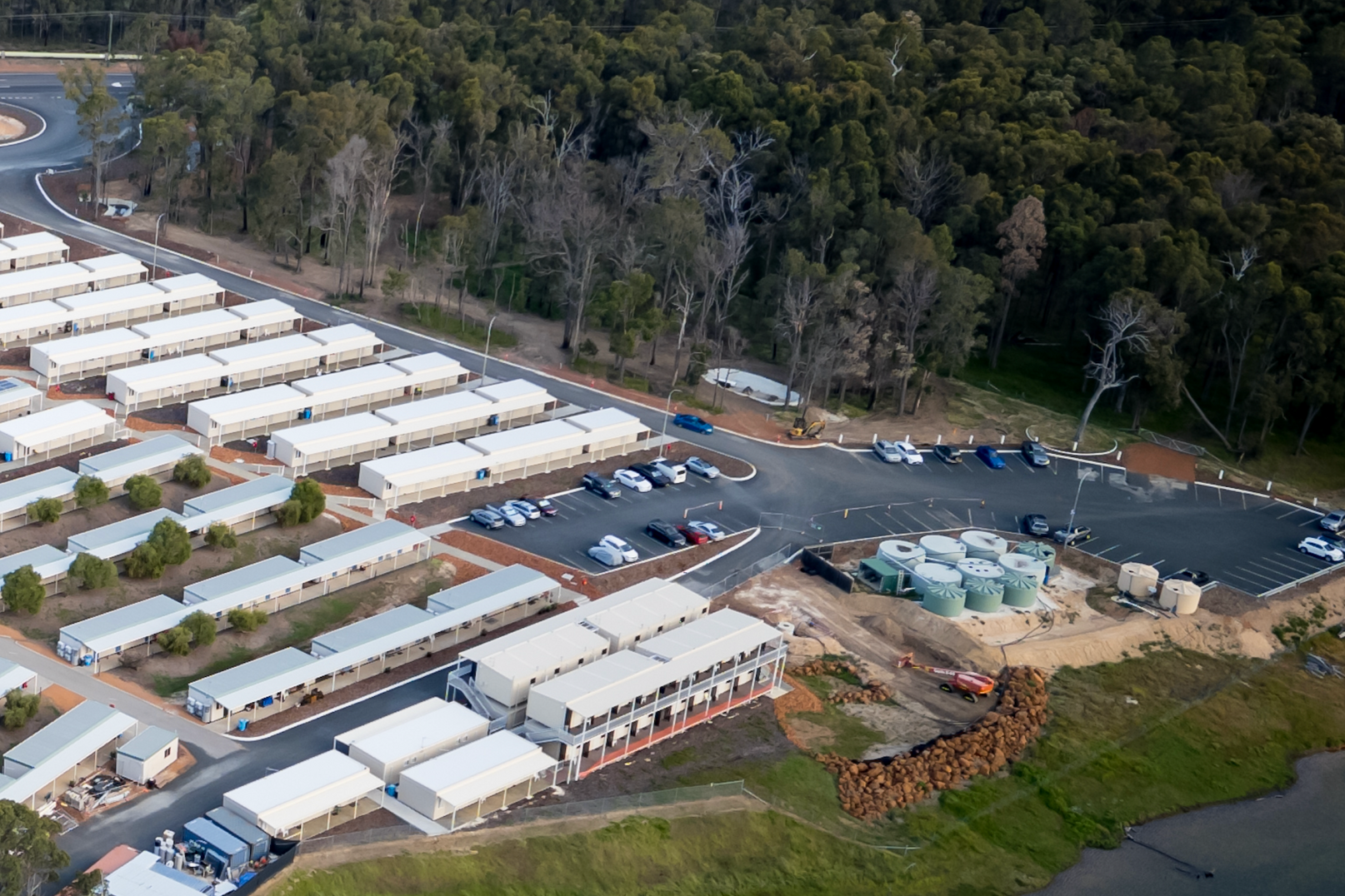 Aerial view of a modular housing complex near a forest and a parking area with vehicles.