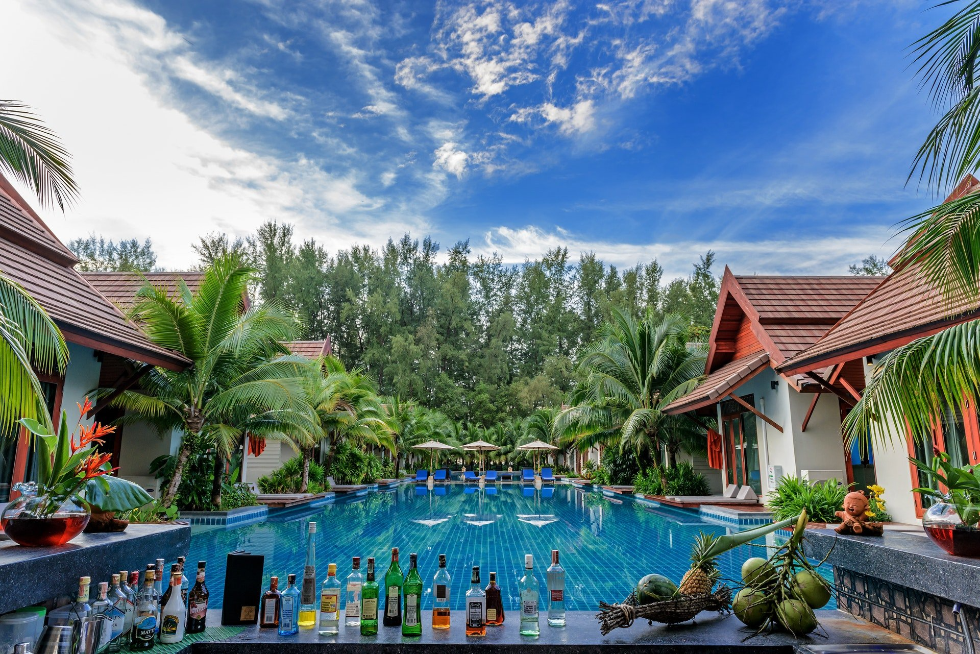 Bar de piscina en un resort tropical; cielo azul con nubes, palmeras, cócteles coloridos y edificios de hotel.