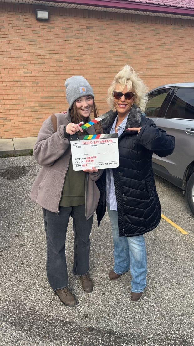Two women are standing next to each other in a parking lot holding a clapper board.
