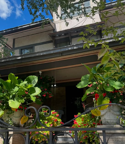 Front porch with lush potted flowers and a dark doorway beneath a balcony