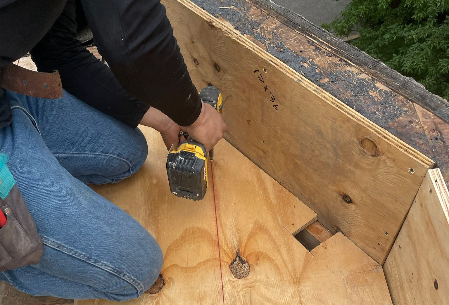 Worker drilling into plywood at a rooftop corner beside a wood-framed wall