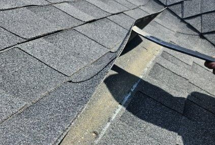 Damaged roof shingles lifted by worker exposing underlayment on a gray shingled roof