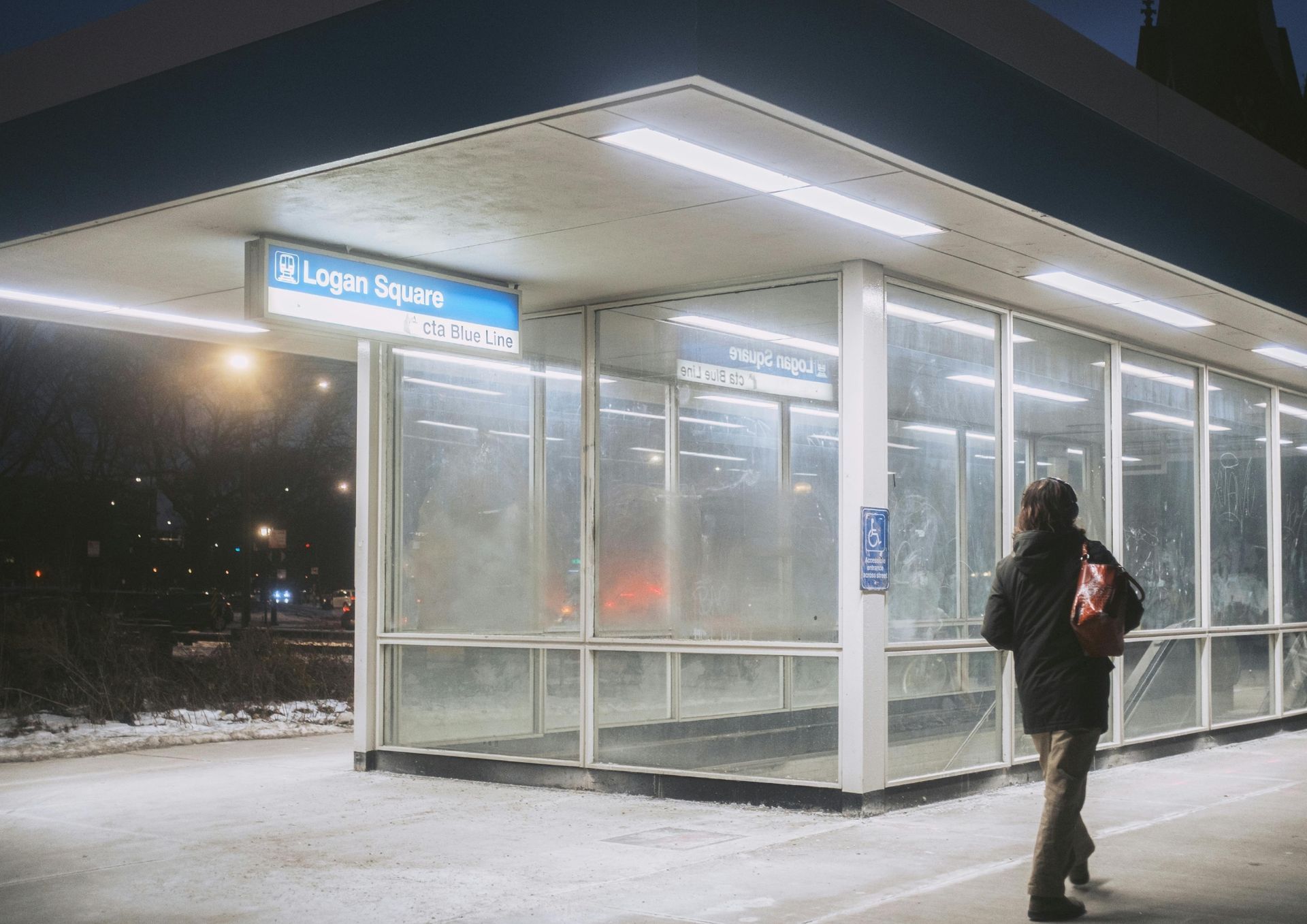 Bus stop at night with a lit shelter and a person standing nearby carrying a bag