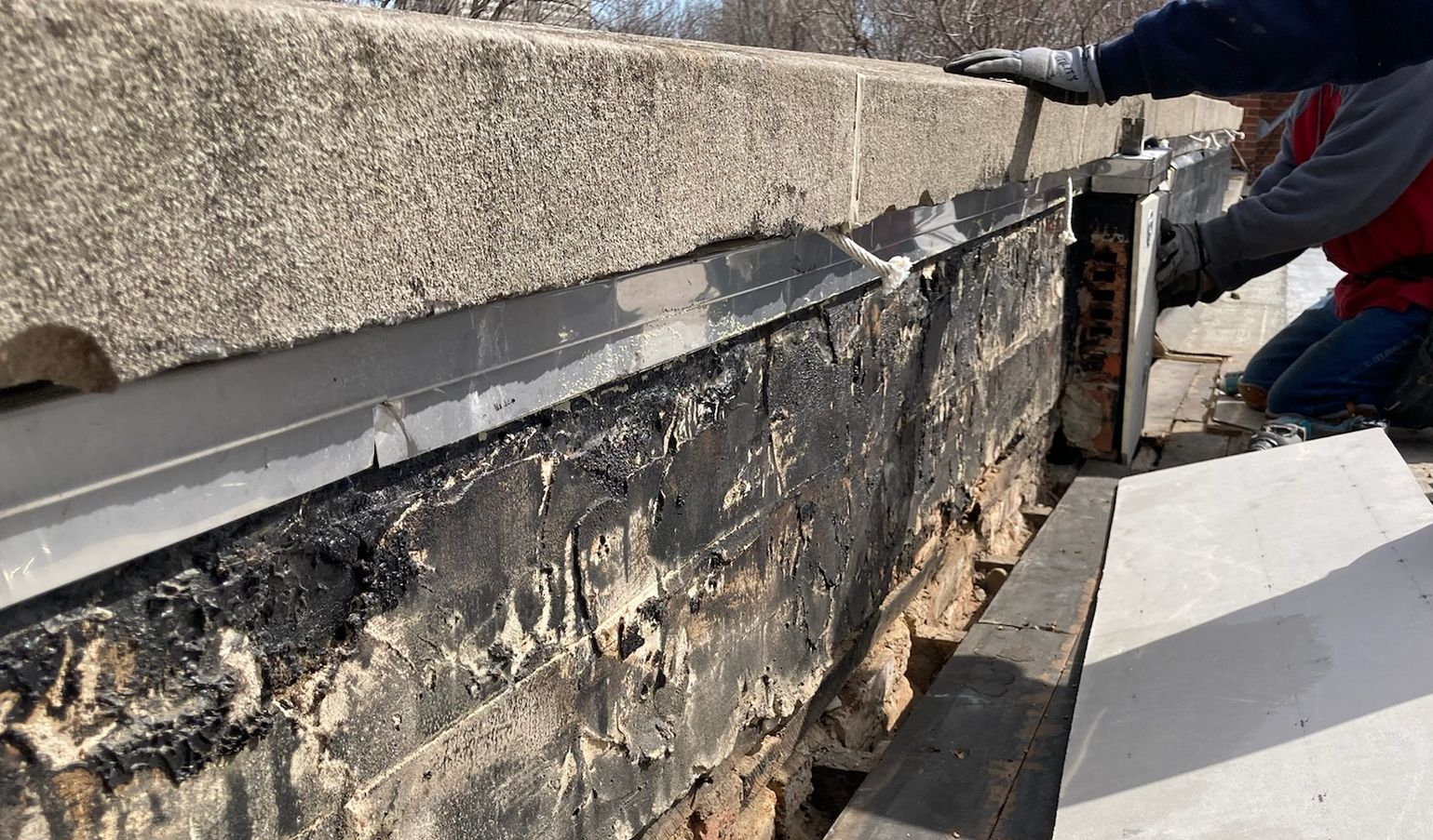 Roofer repairing a damaged roof edge beside a gray parapet wall