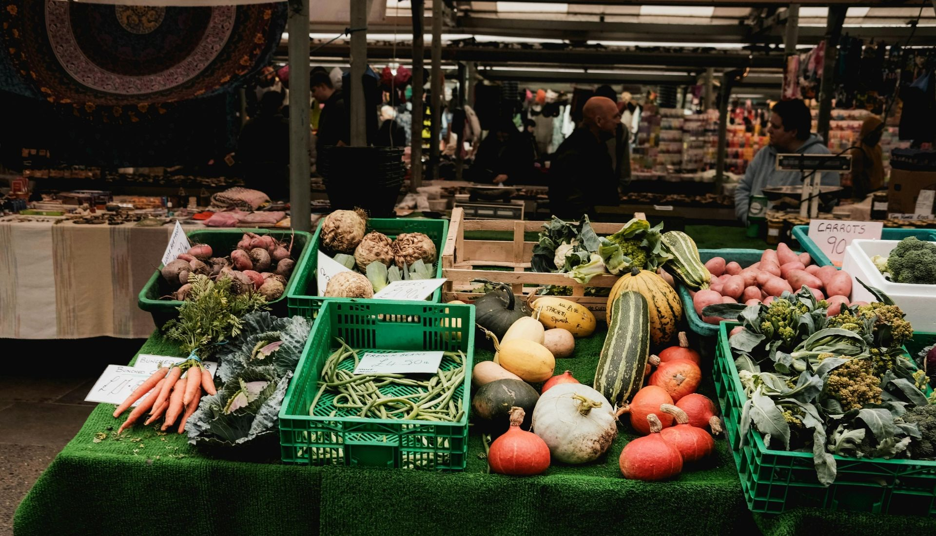 Produce of vegetables and fruits displayed at a market stall with shoppers in the background