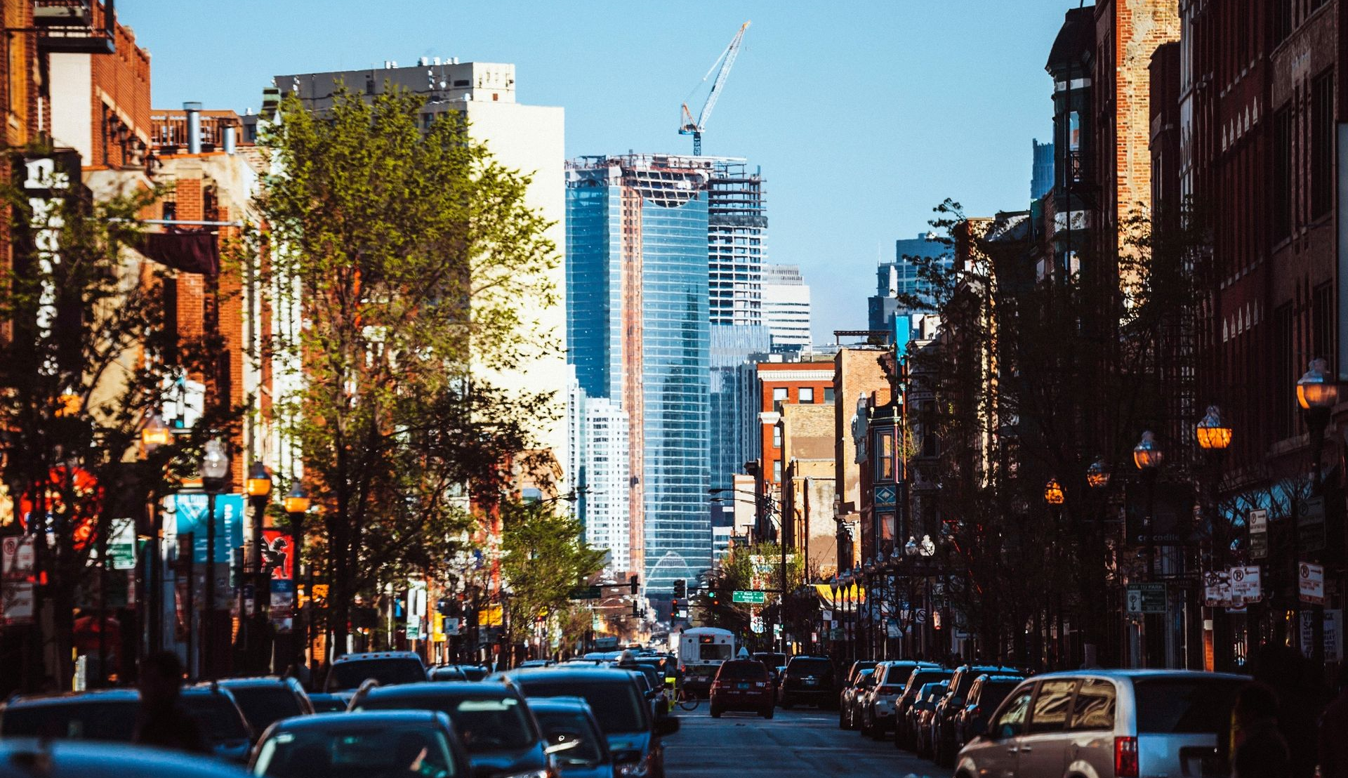 Busy city street with cars, brick buildings, trees, and a blue skyscraper in the distance