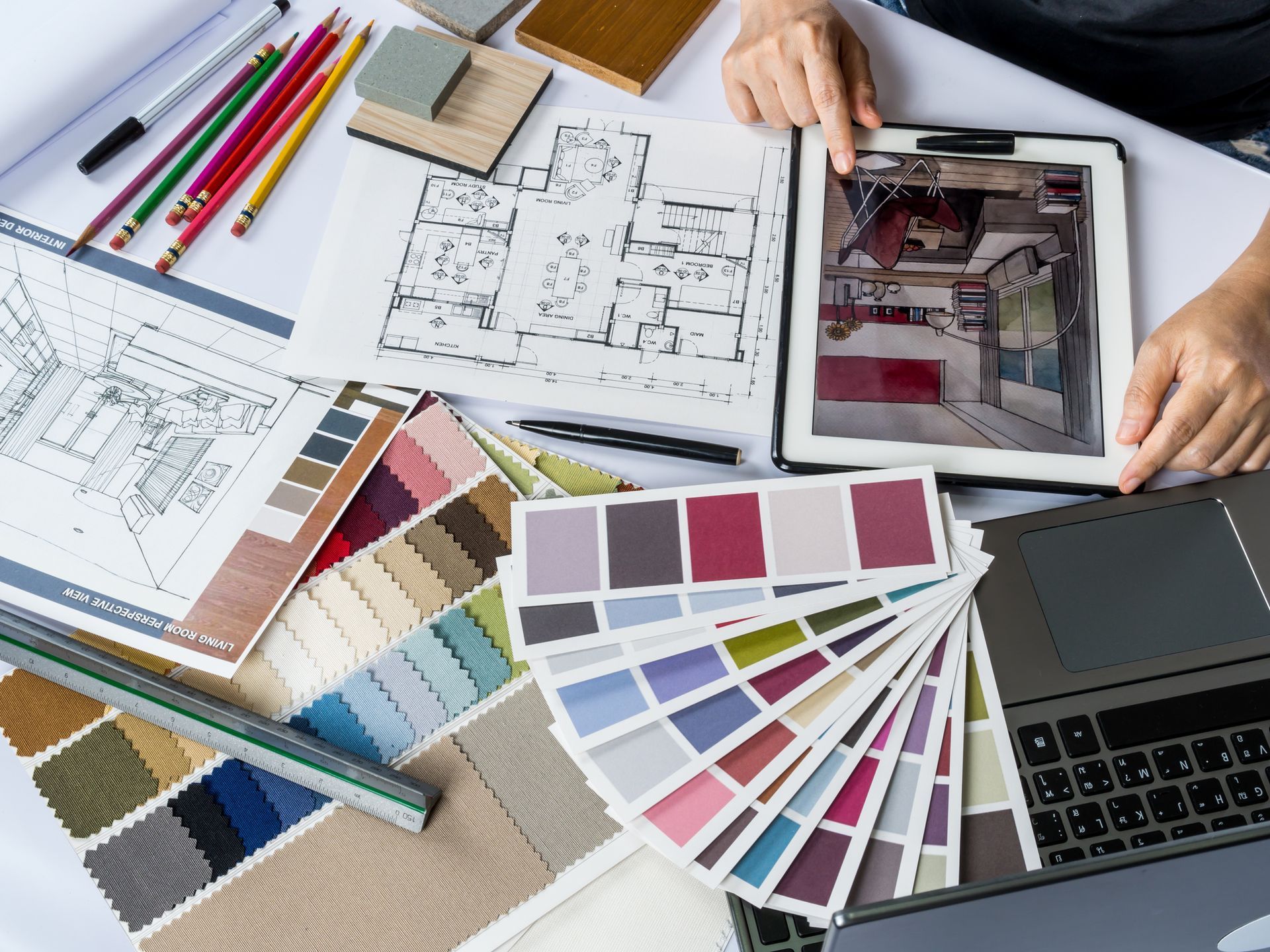 Hands working on interior design tools: floor plans, color swatches, laptop, tablet, and fabrics.
